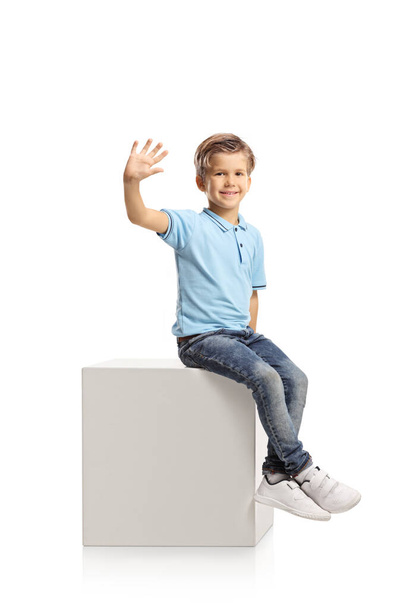Smiling boy sitting on a white cube and waving at camera isolated on white background - Fotografie, Obrázek