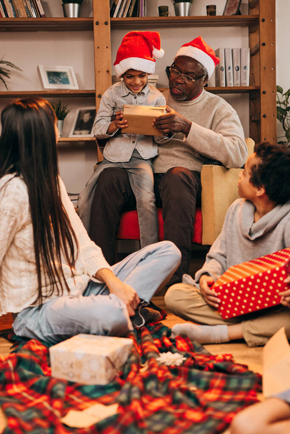 Multikulturní rodina tráví čas spolu na Štědrý večer. Dědeček drží svého vnuka v klíně a dává mu dárek, zatímco šťastná rodina si užívá večer. - Fotografie, Obrázek