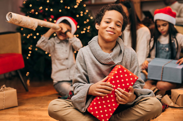 Un garçon afro-américain heureux s'assoit sur le sol à la maison avec un cadeau de Noël dans les mains et regarde la caméra. Dans le fond flou est ses cadeaux d'ouverture de la famille. - Photo, image