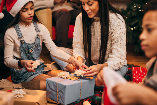 Hija y madre sentadas en el suelo en casa y decorando regalos de Navidad el día de Navidad. - Foto, imagen