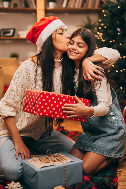 Een liefhebbende moeder kust haar dochter terwijl ze op kerstavond voor de kerstboom op de grond zit. - Foto, afbeelding