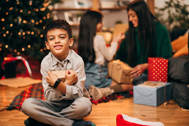Ein junges, glückliches Mädchen mit Weihnachtsmütze auf dem Kopf sitzt an Heiligabend auf dem Fußboden und hält ein Weihnachtsgeschenk in der Hand. - Foto, Bild