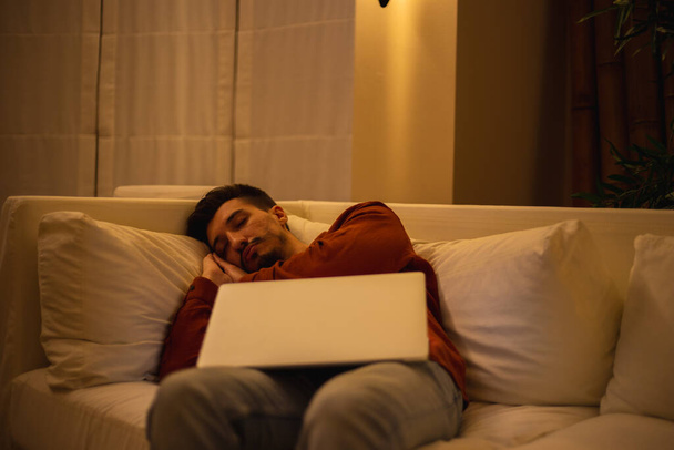 Un jeune homme avec une barbe en chemise rouge travaille avec un ordinateur portable et s'assoit sur le canapé le soir dans la maison. - Photo, image