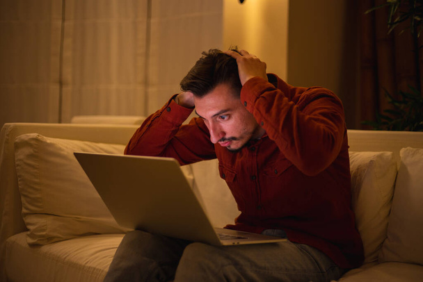 Młody mężczyzna z brodą w czerwonej koszuli pracuje z laptopem i wieczorem siedzi na kanapie w domu.. - Zdjęcie, obraz