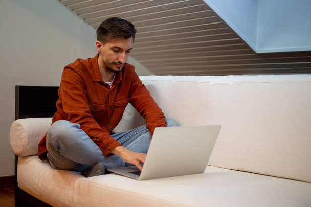 A young man with a beard in a red shirt works with a laptop and sits on the couch in the evening in the house. - Foto, imagen