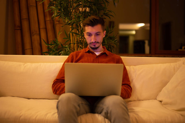 A young man with a beard in a red shirt works with a laptop and sits on the couch in the evening in the house. - Foto, Bild