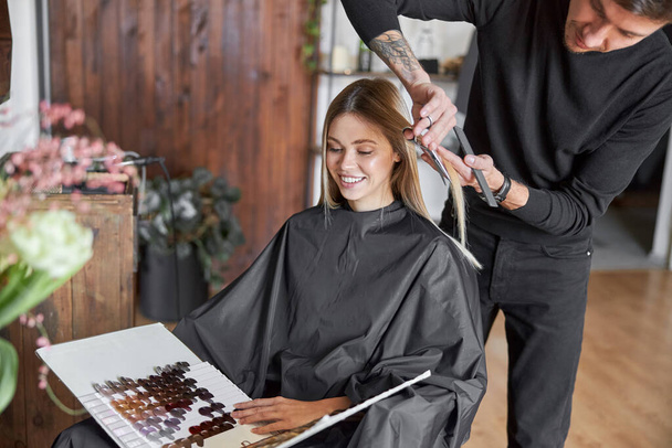 Loira cliente feminina está escolhendo cor no livro, enquanto cabeleireiro está cortando o cabelo - Foto, Imagem