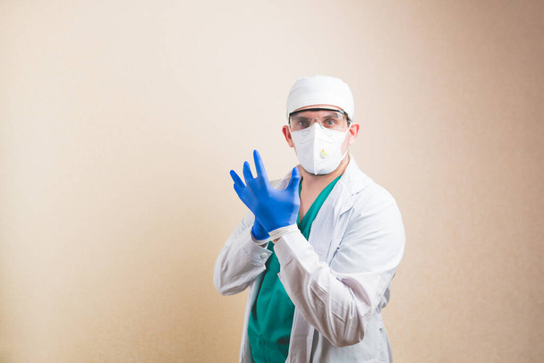 Молодой доктор в белом костюме и защитной дыхательной маске надевает перчатки на руки на желтом фоне изолированы. - Фото, изображение