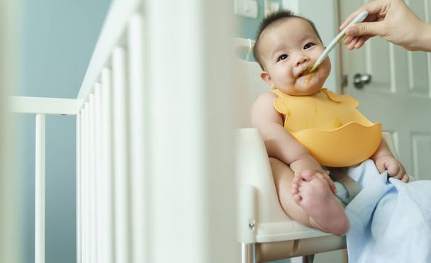 Asian little baby boy wearing yellow bib eating blend food on high chair at home. - Foto, afbeelding