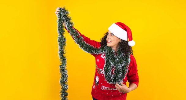 Portrait of hispanic young adult woman with christmas sweater and santa hat on a yellow background in Mexico Latin America - Фото, изображение