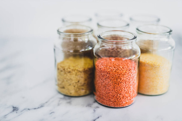 healthy whole grains and legumes in clear pantry jars on marble background including quinoa rice buckwheat lentils and barley, simple vegan pantry ingredients concept - Photo, image