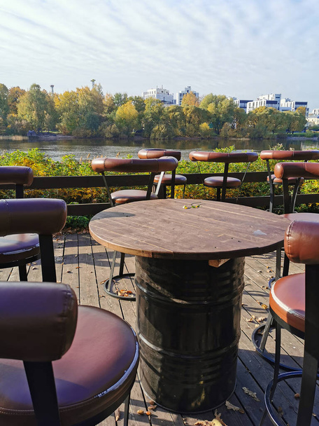 Opuszczona uliczna kawiarnia z wysokimi krzesłami i stołami z metalowych beczek na brzegu rzeki w jesiennym parku na Elagin Island w Sankt Petersburgu. - Zdjęcie, obraz