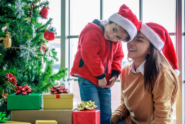 Feliz familia asiática mamá e hija en rojo y blanco Santa Claus sombrero y suéter sonriendo jugando juntos cerca de pino de Navidad con artículos de decoración de brillo y regalo cajas de regalo en la noche de Navidad. - Foto, imagen