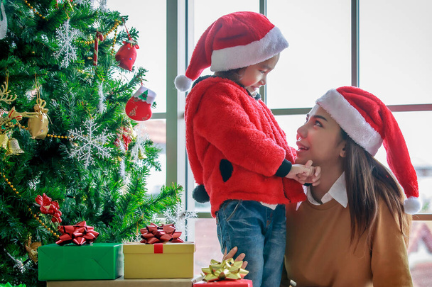 Ευτυχισμένη ασιατική οικογένεια μαμά και κόρη σε κόκκινο και άσπρο καπέλο και πουλόβερ Άγιος Βασίλης χαμογελώντας παίζοντας μαζί κοντά σε χριστουγεννιάτικο πεύκο με glitter διακόσμηση στοιχεία και παρουσιάζουν κουτιά δώρων σε Χριστούγεννα βράδυ. - Φωτογραφία, εικόνα