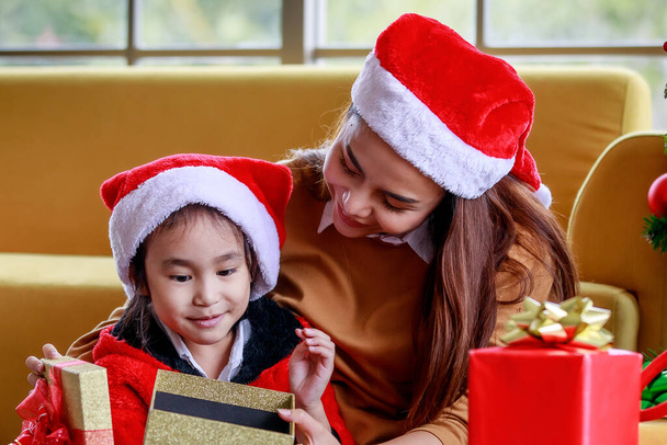 Boldog ázsiai család lánya lány visel pulóver piros és fehér Mikulás kalap ül anya meglepő izgalmas, amikor nyitott ajándék doboz ünnepelni Karácsony este közel karácsonyfa fenyő a nappaliban. - Fotó, kép