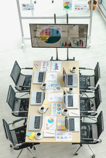 Draufsicht auf Konferenzraum im Büro voller Laptop-Notebooks auf Arbeitstisch mit schwarzen Stühlen und großem Bildschirm und Glaswand mit Post-it-Dokumenten. - Foto, Bild