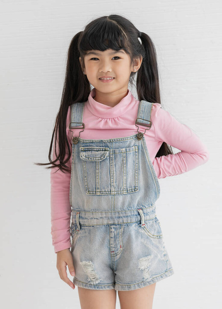 Portrat closeup studio shot of Asian happy little cute preschooler girl with pigtail hair wearing pink long sleeve shirt with jeans overalls standing posing smiling look at camera on white background. - Photo, Image