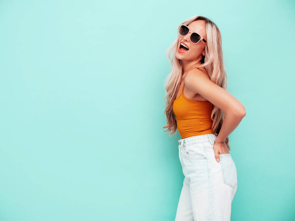 Portrait of young beautiful smiling female in trendy summer clothes. Sexy carefree blond woman posing near blue wall in studio. Positive model having fun indoors. Cheerful and happy. In sunglasses - Foto, immagini
