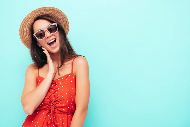 Young beautiful smiling female in trendy summer red dress. Sexy carefree woman posing near blue wall in studio. Positive brunette model having fun in hat and sunglasses - Foto, afbeelding