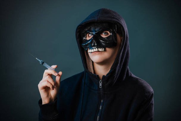 Drug addiction concept. Fear of vaccinations and injections. A person in a sweatshirt with a hood and a skull mask holds a syringe in his hand isolated on a dark background. - Foto, Imagen