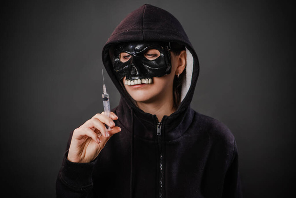 Drug addiction concept. Fear of vaccinations and injections. A person in a sweatshirt with a hood and a skull mask holds a syringe in his hand isolated on a dark background. - Photo, Image
