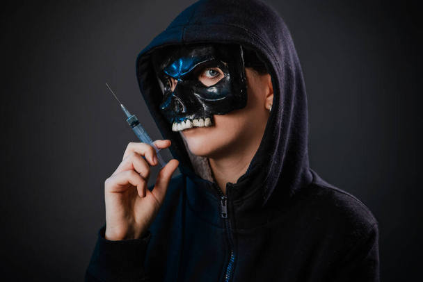 Drug addiction concept. Fear of vaccinations and injections. A person in a sweatshirt with a hood and a skull mask holds a syringe in his hand isolated on a dark background. - Foto, immagini