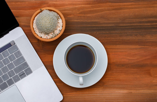 Draufsicht auf Laptop-Notebook-Computer mit schwarzem Kaffee in weißer Keramiktasse und Kaktuskanne auf Holzhintergrund mit Kopierraum. - Foto, Bild