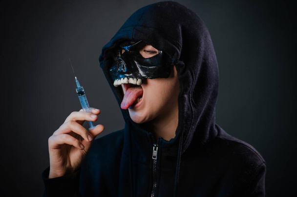 Drug addiction concept. Fear of vaccinations and injections. A person in a sweatshirt with a hood and a skull mask holds a syringe in his hand isolated on a dark background. - Fotoğraf, Görsel