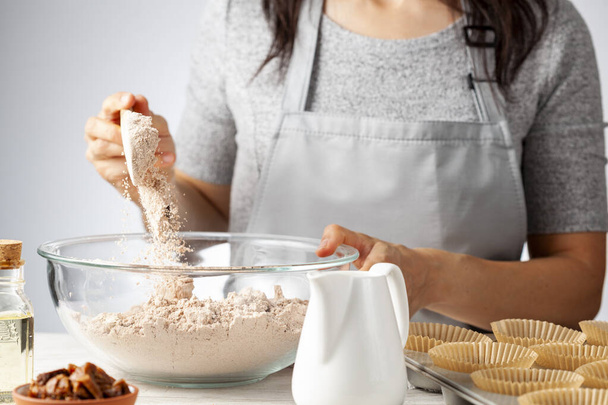 A woman is adding ingredients for making delicious fruit and nut cupcake on white marble countertop background. Muffin tin with liner, ingredients, and utensils are seen. Caucasian woman wearing apron - Photo, Image
