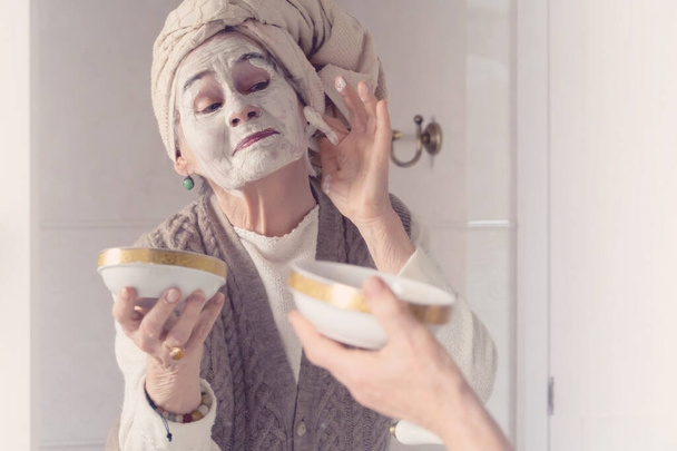 літня жінка з рушником на голові посміхається і наносить маску для очищення
 . - Фото, зображення