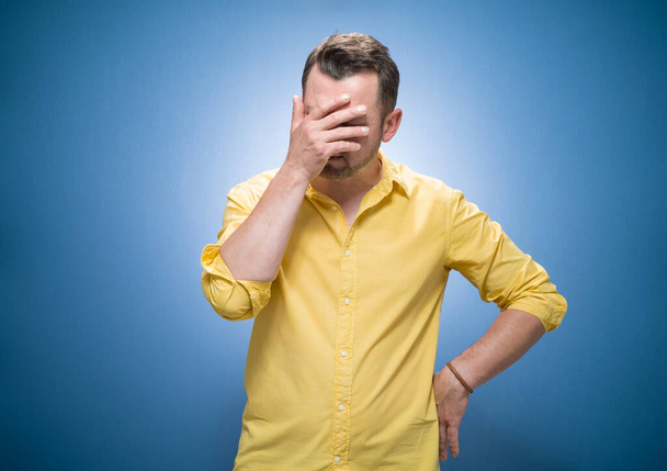 Young man covering face with hand. Shame over blue background, dresses in yellow shirt. Ashamed guy holding hand on her head. Studio shot - Fotoğraf, Görsel