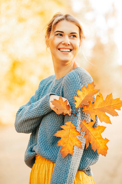 Porträt einer jungen Frau mit Herbstlaub in der Hand, die im herbstlichen Wald oder auf der Straße einen blauen Strickpullover trägt und glücklich lächelt, nach einem anstrengenden Arbeitstag allein zu sein..  - Foto, Bild