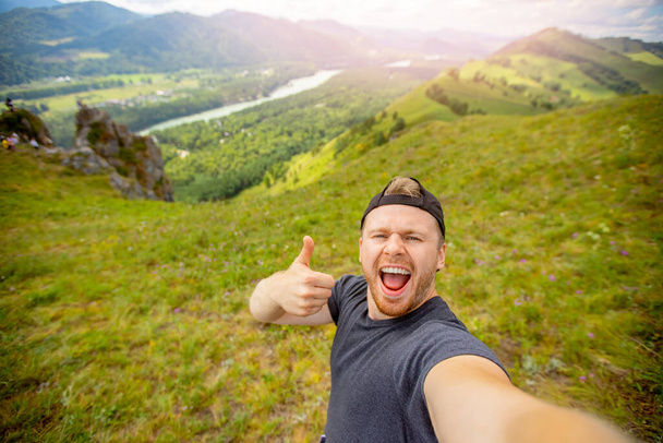 Happy young man blogger on background of mountains in summer sunset. Lifestyle Russia Altay traveler makes selfie photo - Photo, image