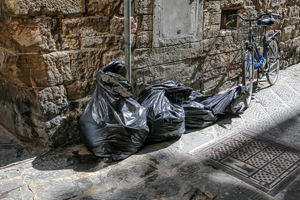 Sacs poubelles en plastique noir et un vieux vélo dans une ruelle arrière par un vieux mur de pierre texturé et un trou d'homme - Photo, image