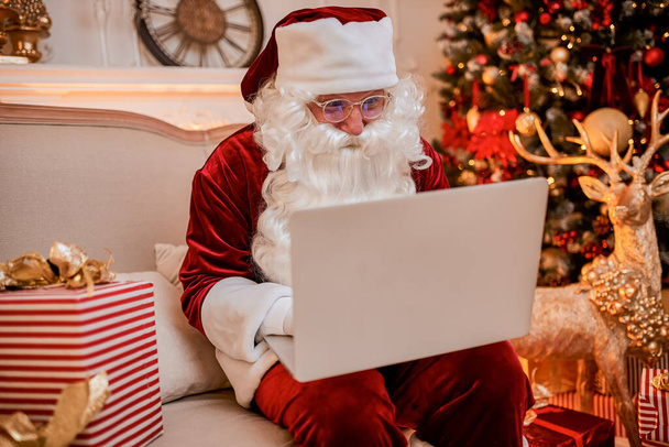 Mikulás ül az otthonában, és e-mailt olvas a laptopon a karácsonyi kérő vagy kívánságlista közelében a kandalló és a fa ajándékokkal. Újév és Boldog Karácsonyt!  - Fotó, kép