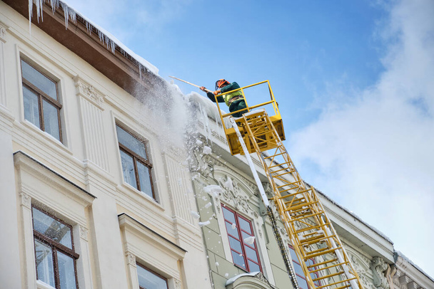 Public utilities on special vehicles remove icicles from the roofs of houses on the Rynok square in Lviv. Roof Winter Workers. Large icicles hanging from the roof - Photo, Image