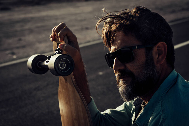 Ciemny cień i jasny portret przystojnego dorosłego mężczyzny z brodą i okularami przeciwsłonecznymi trzymającego długą deskę z asfaltową drogą w tle. - Zdjęcie, obraz