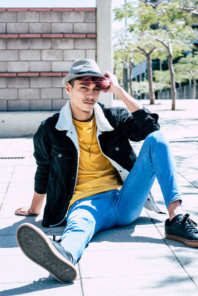 Young trendy teenager sitting on the asphalt ground looking at the camera in outdoor leisure activity and relax portrait. - Foto, imagen