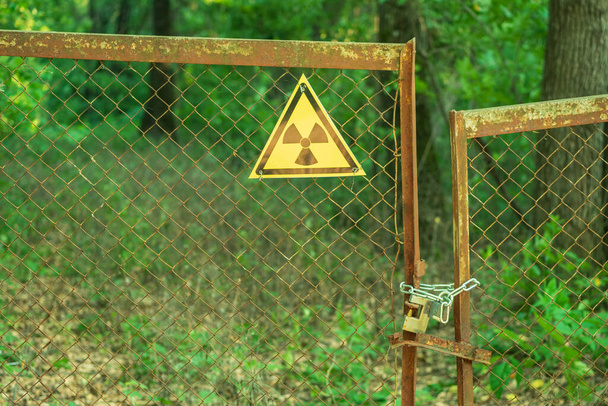 A radiation sign tied with wire to the bars of an iron gate. The flaps of the gate are wrapped with a chain and a lock. In the background are tree trunks and green forest - Zdjęcie, obraz