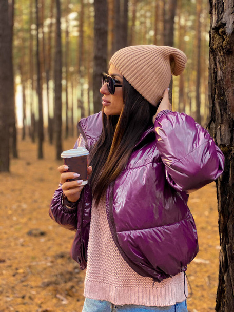 Gelukkige jonge vrouw in gebreide trui en hoed met warme koffie wandeling in het bos - Foto, afbeelding