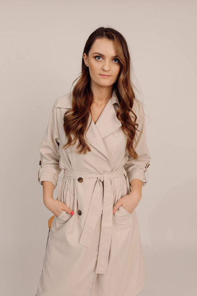 Young beautiful cute serios long brown wavy hair woman top manager in beige trench coat looks at camera, smiles positively, keeps her hands in pockets. Portrait - Foto, Bild