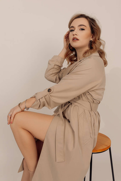 Jovem bela mulher caucasiana com cabelo castanho vestindo trench trench coat moderno sentado na cadeira. Pessoas reais, estúdio filmado em fundo neutro. Roupa elegante sensual para primavera ou outono  - Foto, Imagem