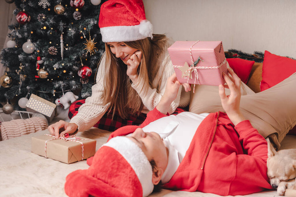 Romanttinen pari vaihtaa lahjoja lähellä koristeltu joulukuusi. Mies antaa joululahjaksi yllätys hymyilevä nainen villapaita rentoutuen sohvalla. Poikaystävä ja tyttöystävä. - Valokuva, kuva