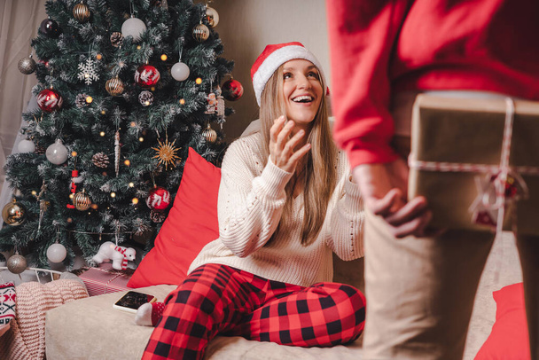 Překvapuje ji... Detailní zadní pohled na muže v červeném svetru drží vánoční dárkovou krabici za zády, zatímco stojí u zdobeného vánočního stromečku. Rodinný pár na vánoční prázdniny. - Fotografie, Obrázek