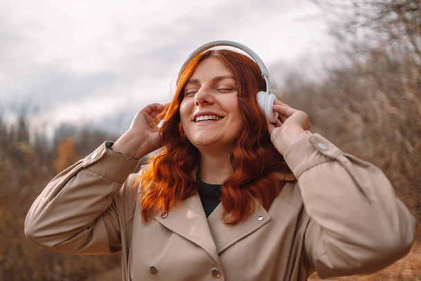 Happy young laughing curly hair woman with closed eyes in oversized beige trench coat listening to music with smartphone and wireless headphones at city park - Photo, Image