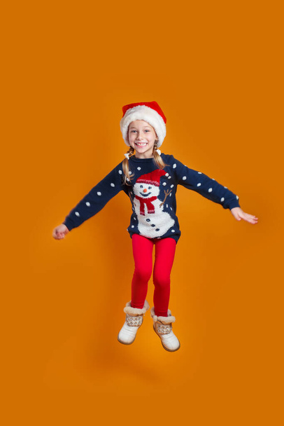 Happy little smiling girl in Santa hat is jumping on orange background. Merry Christmas - Fotoğraf, Görsel