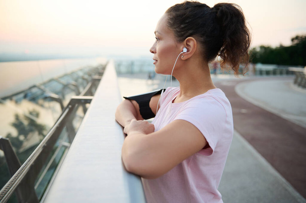 Close-up portrait of confident serene middle aged Hispanic woman with earphones in pink t-shirt enjoying beautiful riverscape at sunrise standing on the city bridge after morning cardio workout - Photo, Image