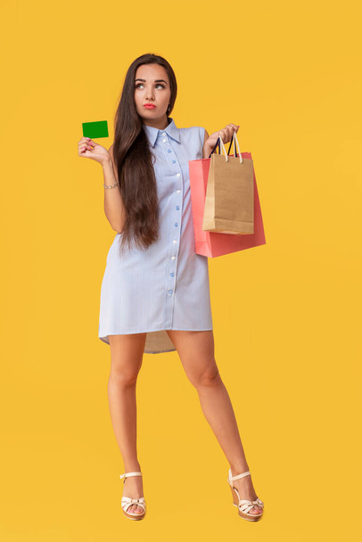 Charmante jolie fille sexy montrant carte de crédit et tenant de nombreux paquets colorés à la main, il est confortable d'utiliser une carte bancaire pour faire du shopping, debout sur fond jaune
 - Photo, image