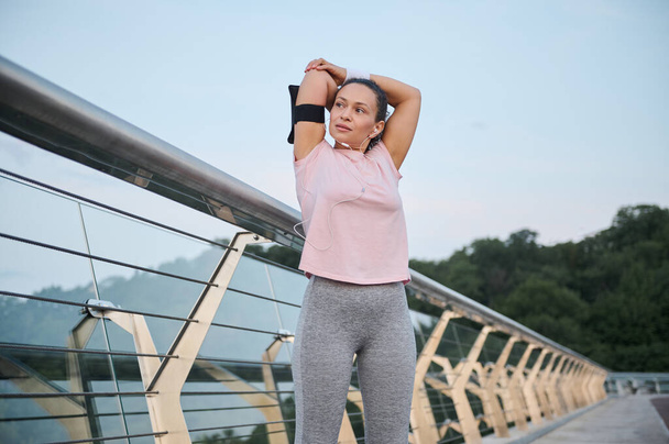 Hermosa mujer, atleta con camiseta rosa estira su cuerpo, de pie en un puente de cristal de la ciudad durante un entrenamiento matutino al amanecer. Deporte, estilo de vida saludable y activo, vitalidad, concepto de pérdida de peso - Foto, imagen