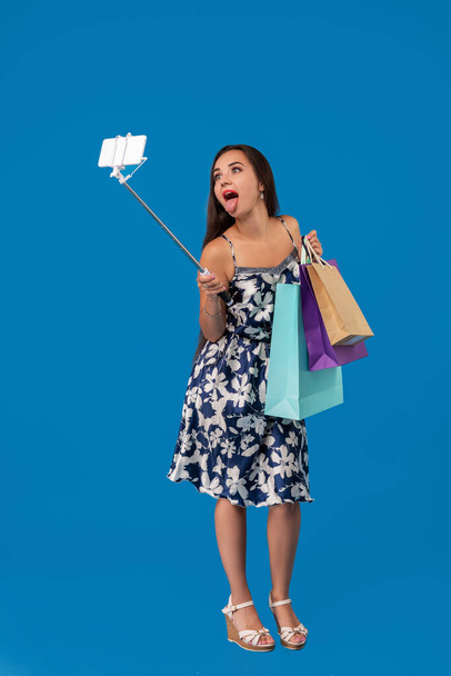Nuori tyylikäs nainen poseeraa ja ottaa selfie puhelimessa ostoskassit sinisellä taustalla
 - Valokuva, kuva
